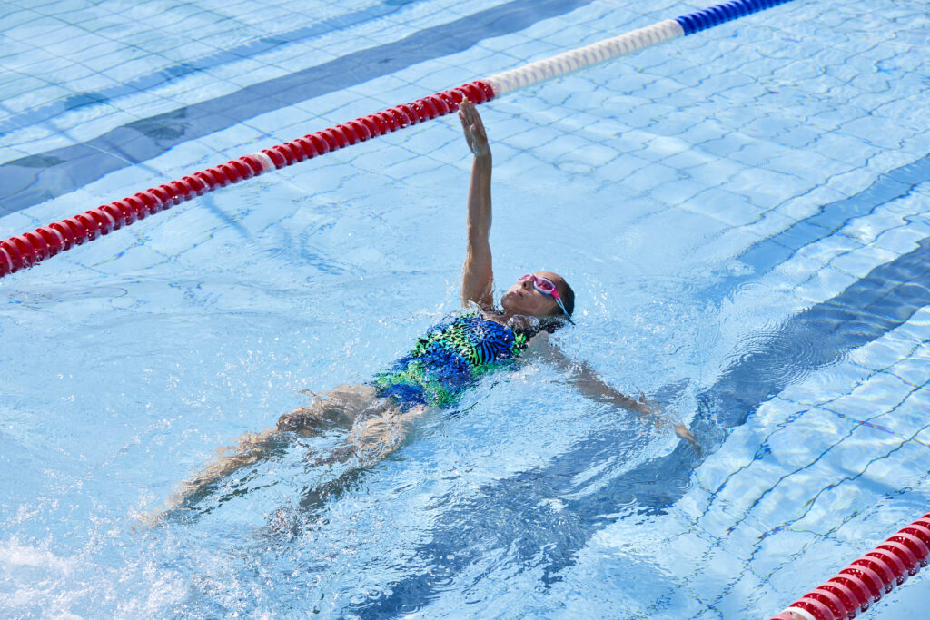 Photo looking downwards upon a young girl swimming backstroke in a swimming pool, with one arm oustretched above water.