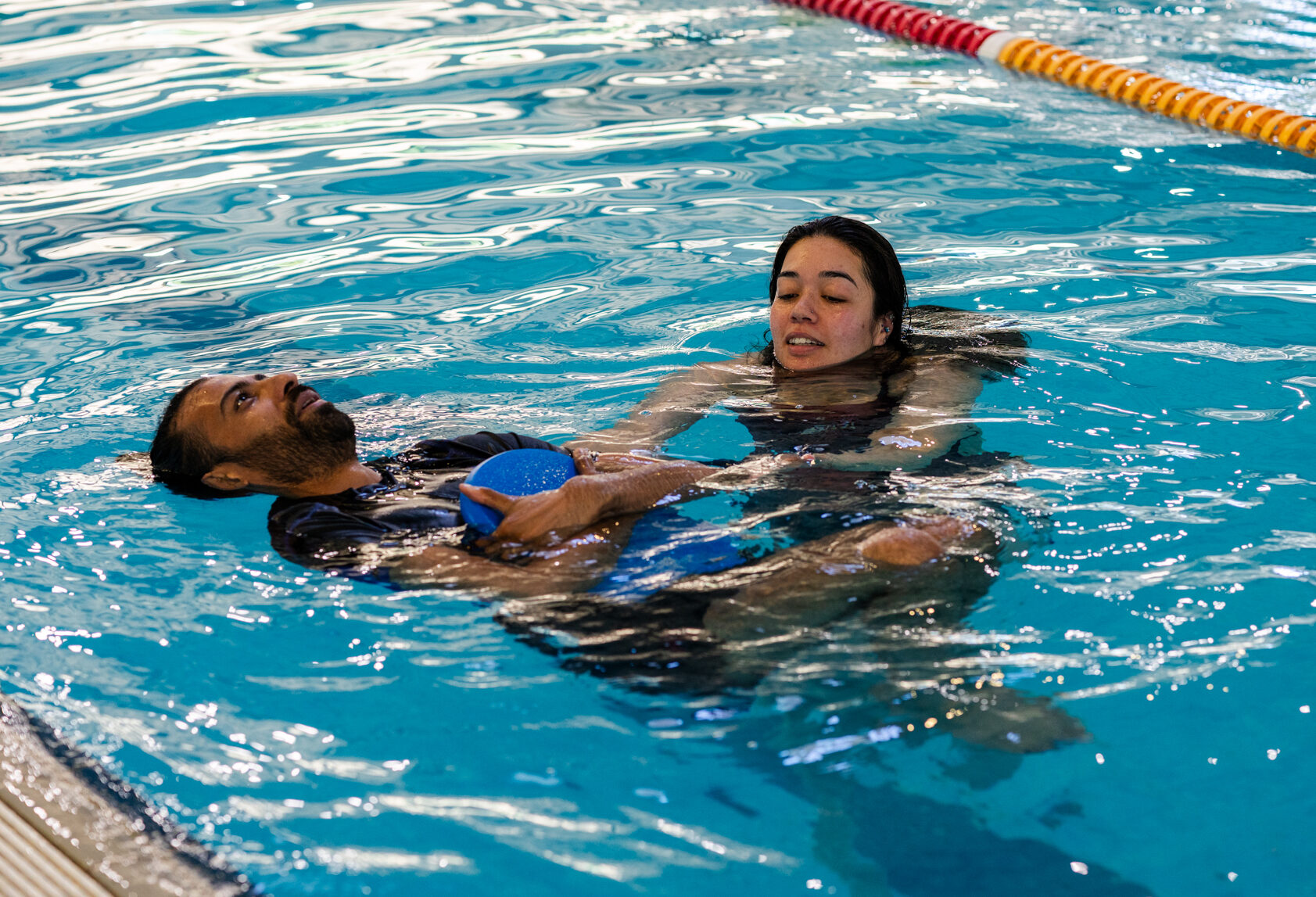 Photo of adult floating in swimming pool on their back whilst holding kickboard beside swim instructor